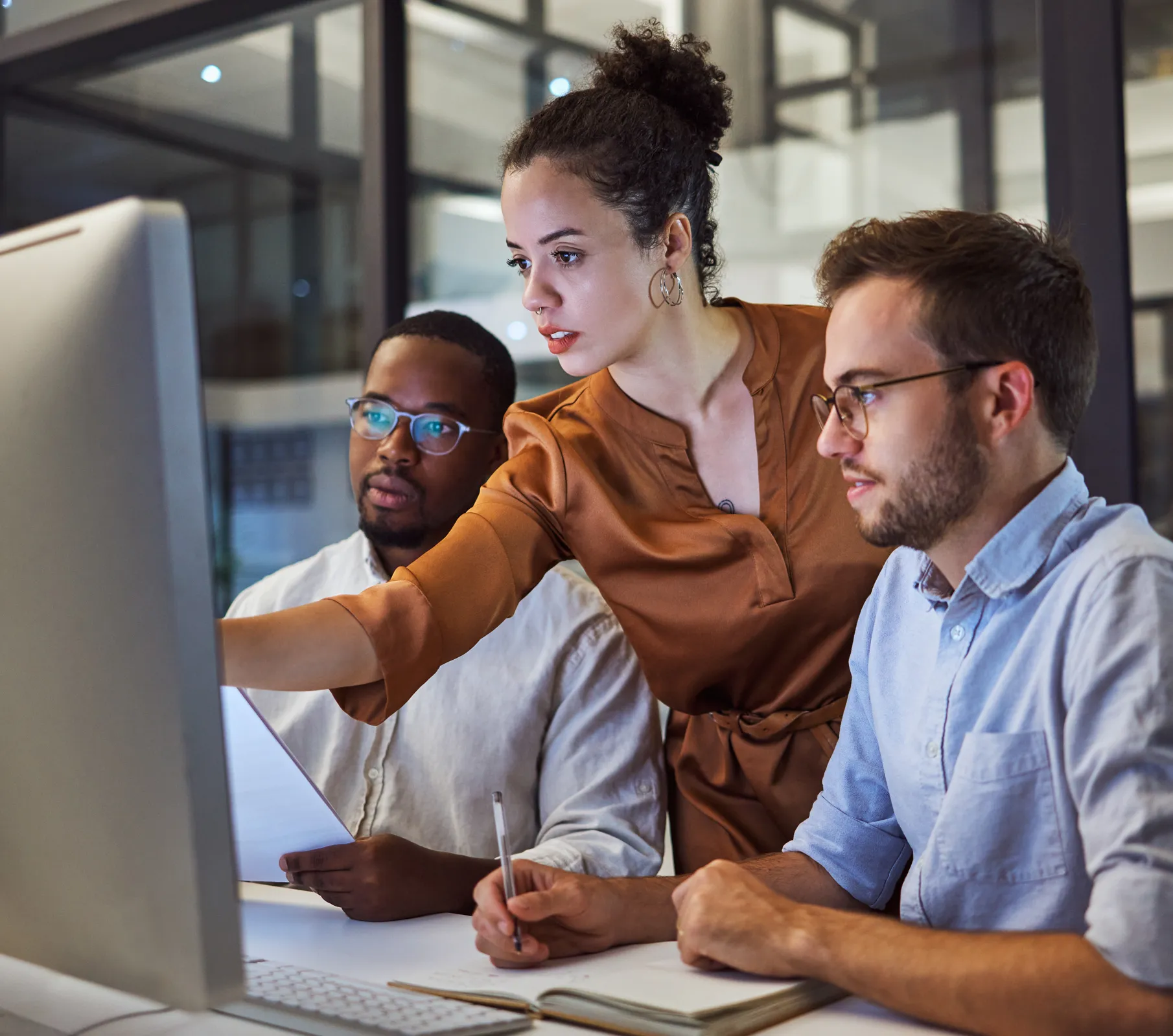 an image of three workers standing around a desktop monitor, with one of them pointing to something on the screen