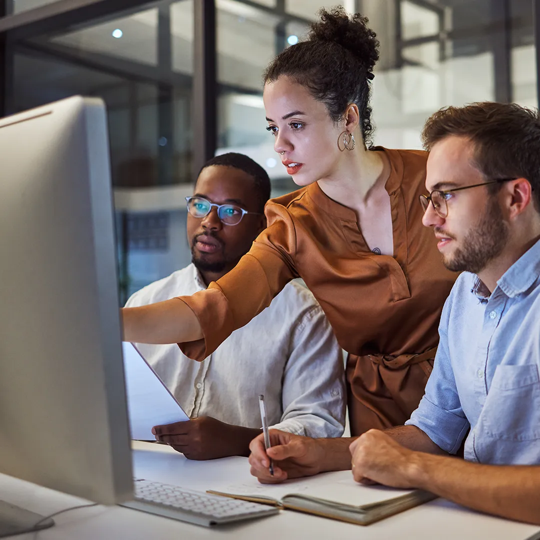 an image of three workers standing around a desktop monitor, with one of them pointing to something on the screen