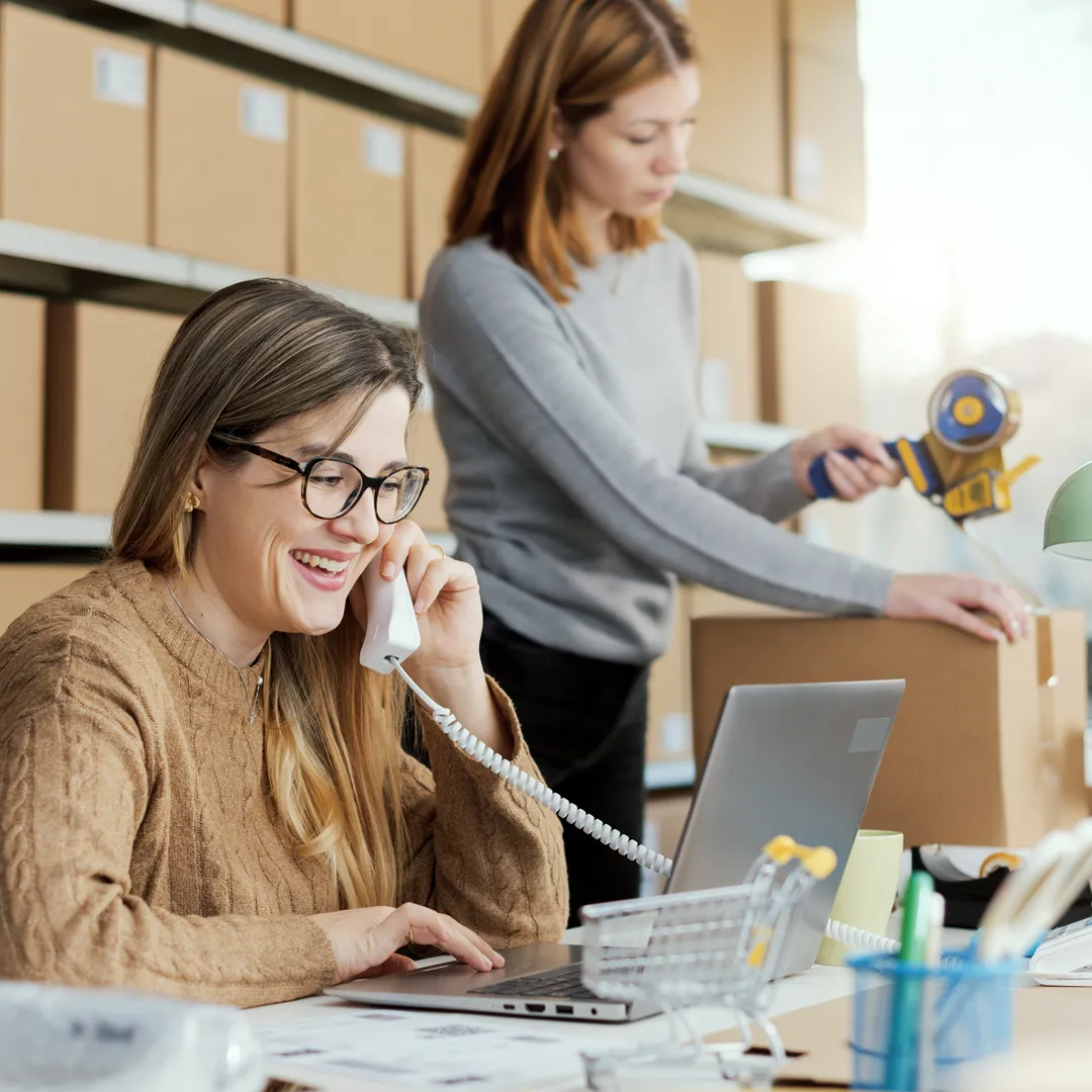 an image of two office workers, one of them on a phone call in front of a tablet, the other is taping a cardboard box