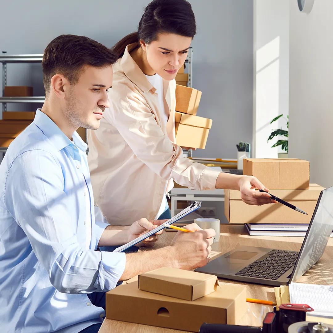 two office workers surrounded by cardboard packaging looking at a laptop